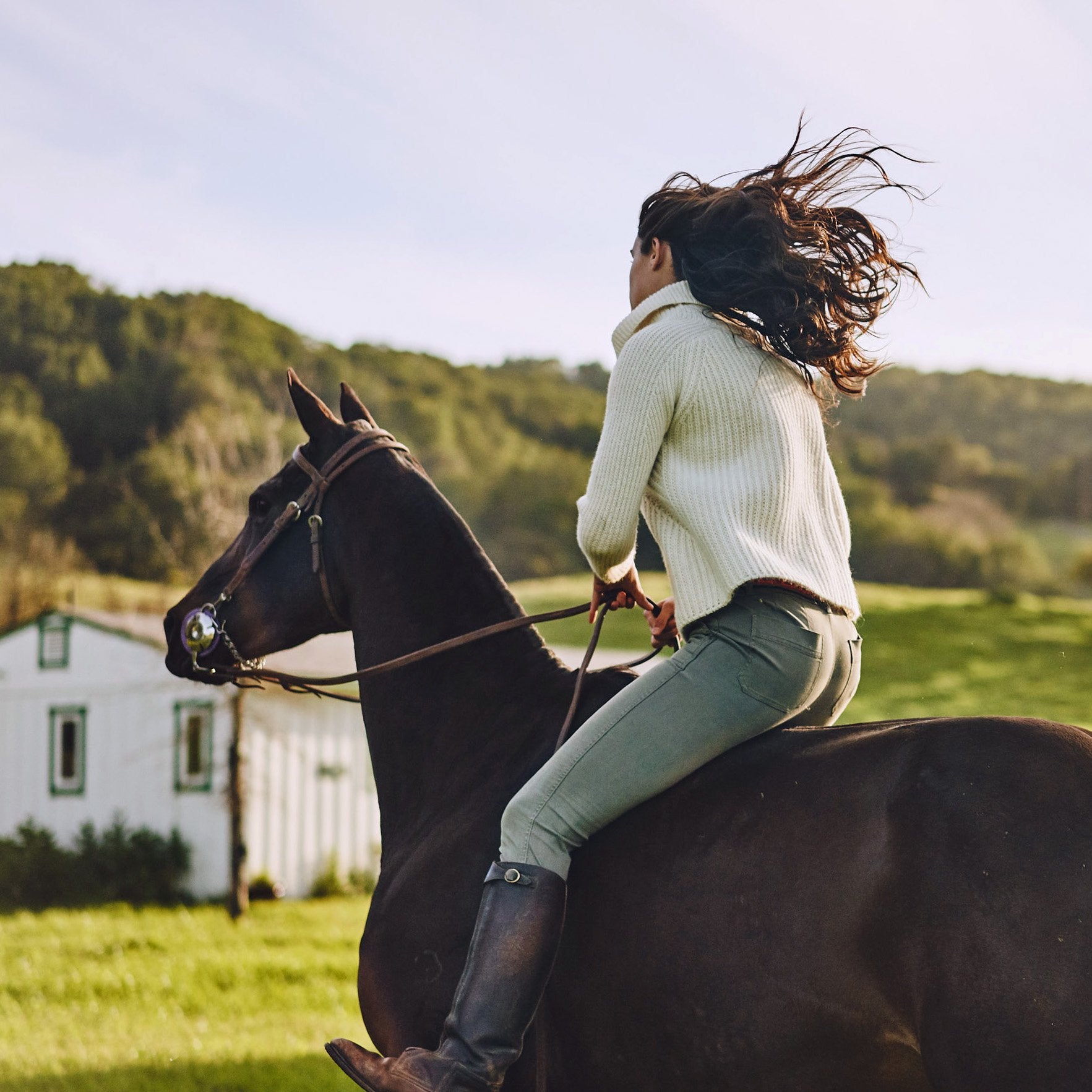 Woman riding horse a horse in the countryside wearing an Alpaca Ribbed Cowl Neck Sweater in Cream by Stick & Ball