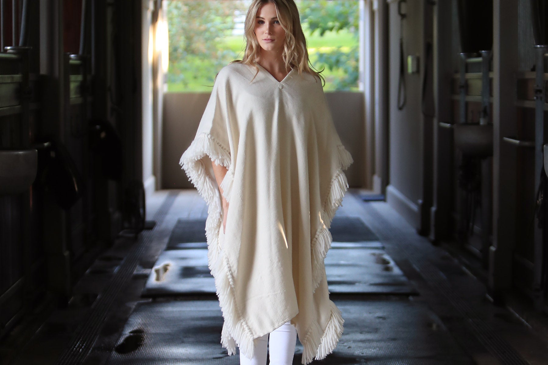 Collection image for New Ponchos and Ruanas - model standing in dark horse barn wearing our long fringed alpaca poncho in winter white
