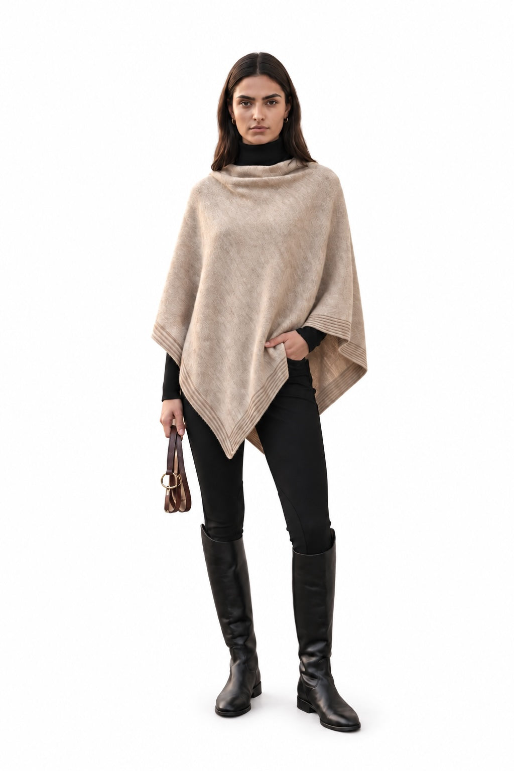 A woman modeling a taupe knit poncho over a black turtleneck, black pants, and black knee-high boots, holding equestrian reins against a plain white background.