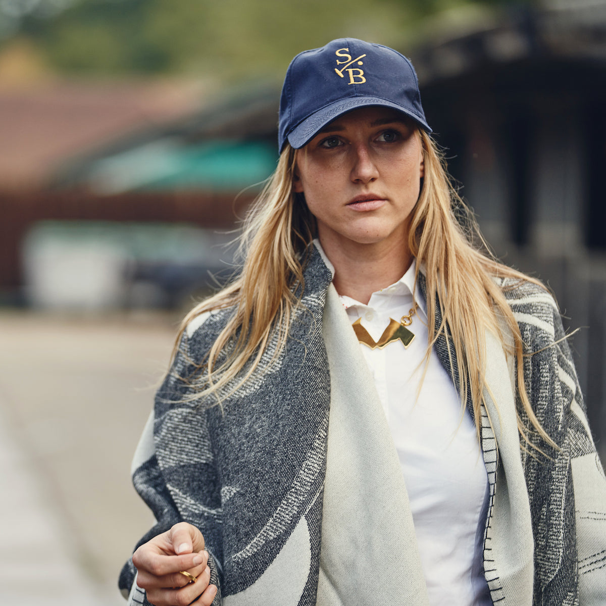 A woman wearing a navy blue Stick & Ball baseball cap with a gold embroidered monogram, a white collared shirt, a gold Kissing Horses Necklace and a gray and white Polo Pony Throw wrap, photographed outdoors with a blurred background.