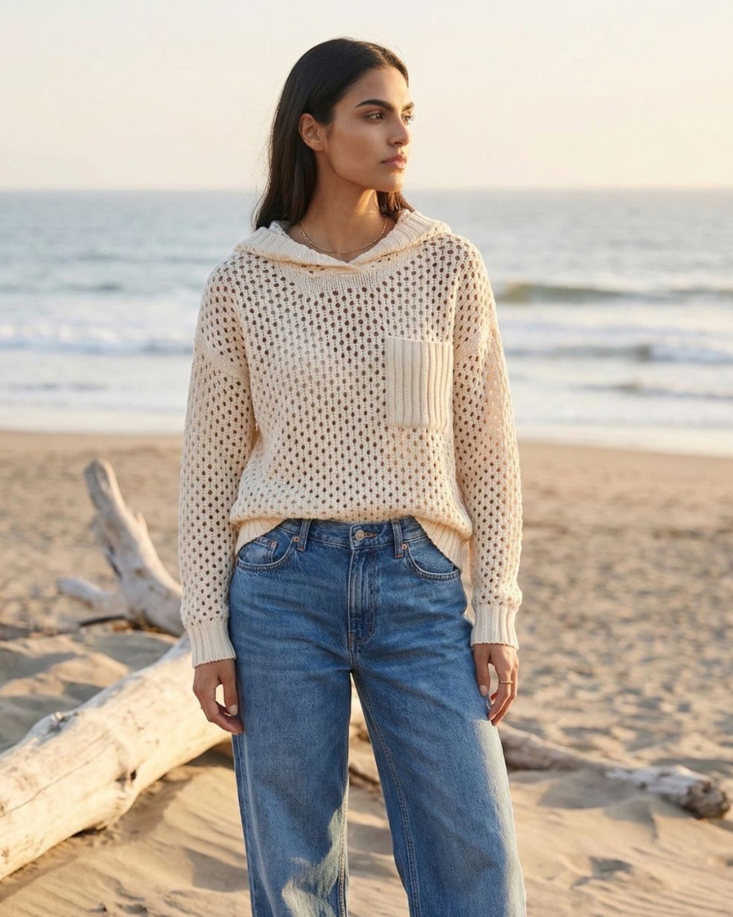 Woman wearing a beige mesh top and blue jeans standing on a beach.
