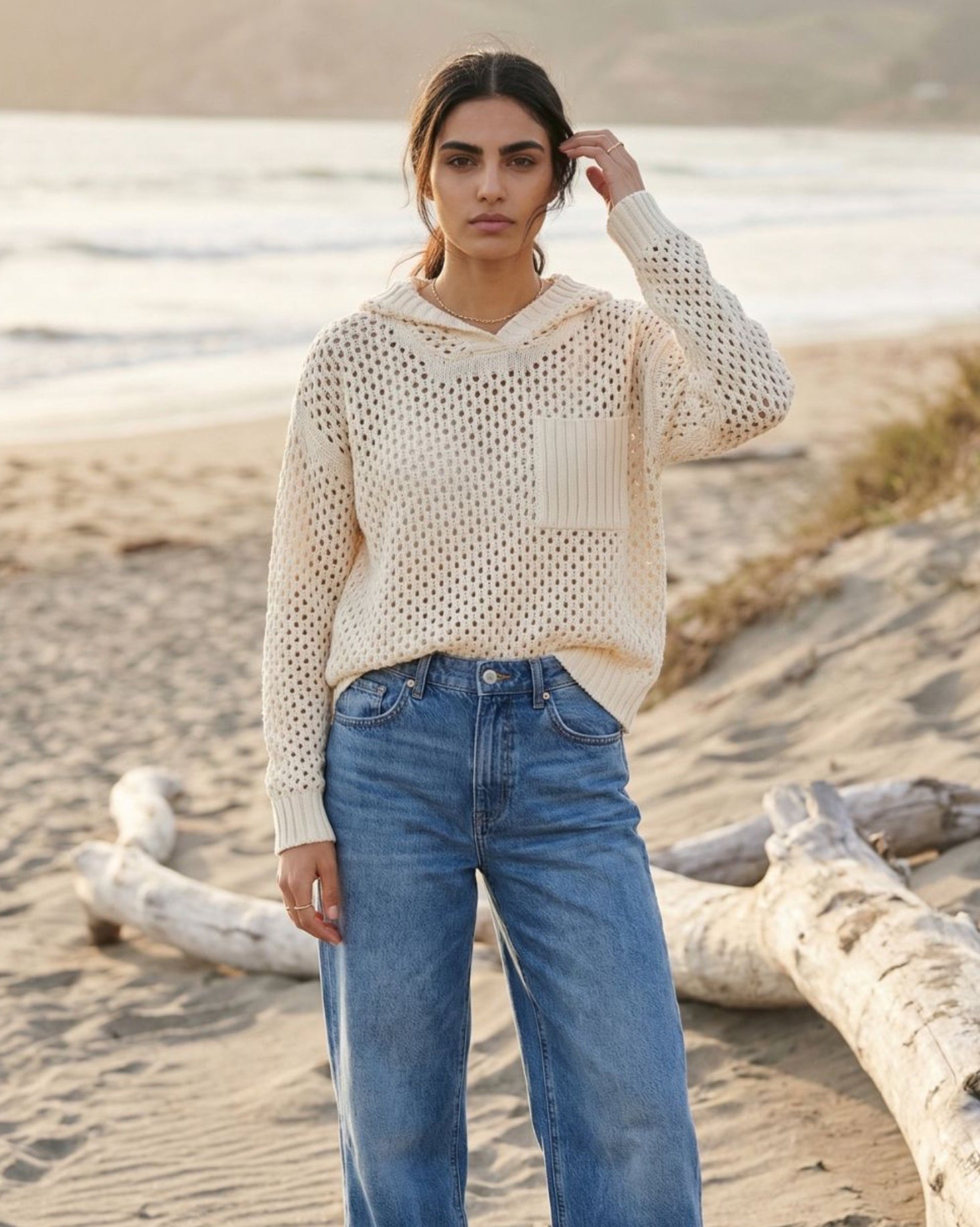 Woman wearing a beige knitted sweater and blue jeans standing on a beach.